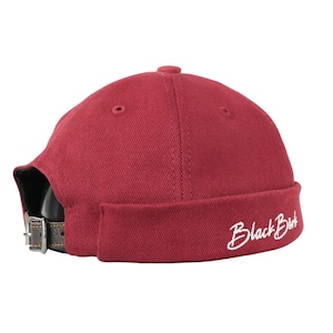 May include: Maroon skull cap with a curved brim and a brown leather strap. The cap has the words "Black Bird" embroidered in white on the side. The cap has a small button on top.