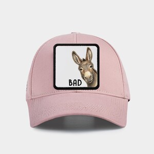 Pode incluir: Boné de beisebol rosa claro com um patch quadrado. O patch apresenta um burro de desenho animado e a palavra "BAD" em preto. O burro é marrom e branco, com a cabeça virada para o lado.