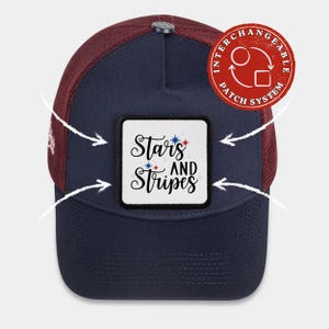 May include: A navy blue trucker cap with a maroon mesh back and a square white patch on the front. The patch features the black text Stars AND Stripes with small red and blue star accents. A red circular badge indicates an interchangeable patch system.