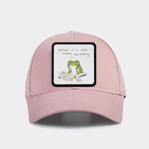May include: A pale pink baseball cap with a square patch. The patch displays a cartoon frog looking thoughtful, with the words "Whatever it is, you're probably over-thinking it." The frog is surrounded by papers and a coffee cup.