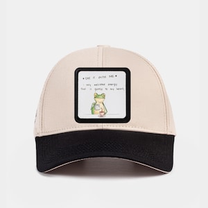 May include: A beige and black baseball cap with a square patch showing a cartoon frog with a cup. The patch has the text "SAY IT WITH ME" and "I only welcome energy that is gentle to my heart."