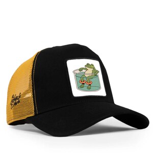 May include: Black and yellow trucker cap with a patch showing a cartoon frog relaxing in a glass of water with orange floatation rings. The cap has a curved peak and a mesh back.
