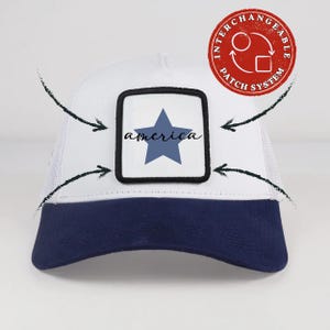 May include: A white trucker cap with a dark blue peak and a square patch on the front. The patch features a blue star and the word america in black script. A red circular logo in the top right corner reads INTERCHANGEABLE PATCH SYSTEM.