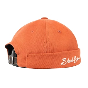 Op de afbeelding: Oranje skullcap met een gebogen klep en een bruine leren riem met een metalen gesp. De pet heeft de tekst "Black Bird" in het wit geborduurd op de klep.