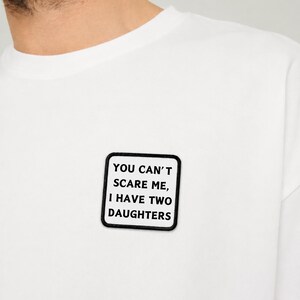Könnte beinhalten: Ein weißes T-Shirt mit einem quadratischen Aufnäher auf der Brust. Der Aufnäher hat einen schwarzen Rand und einen weißen Hintergrund mit dem schwarzen Text: YOU CAN'T SCARE ME, I HAVE TWO DAUGHTERS. Das T-Shirt hat einen Rundhalsausschnitt.