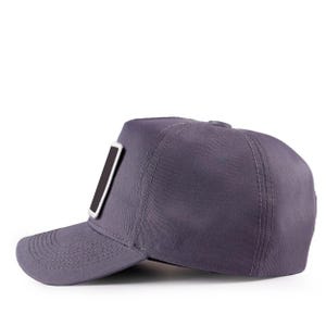 May include: A solid dark grey baseball cap featuring a structured crown and a curved brim. The cap includes a rectangular patch on the front panel with a black centre and a thick white border. The design shows visible stitching along the brim.