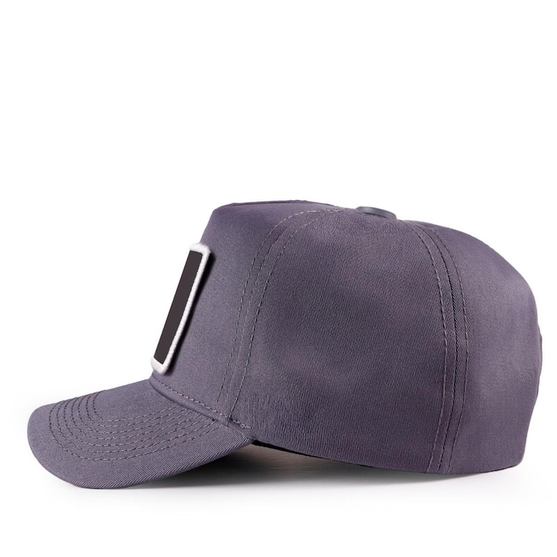 May include: A solid dark grey baseball cap featuring a structured crown and a curved brim. The cap includes a rectangular patch on the front panel with a black centre and a thick white border. The design shows visible stitching along the brim.