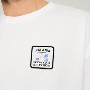 Puede incluir: Una camiseta blanca con un parche cuadrado de borde negro. El parche muestra ilustraciones de tres envases de leche y el texto en negro: JUST A DAD WHO ALWAYS CAME BACK WITH THE MILK, con dos líneas horizontales a cada lado de la última línea.