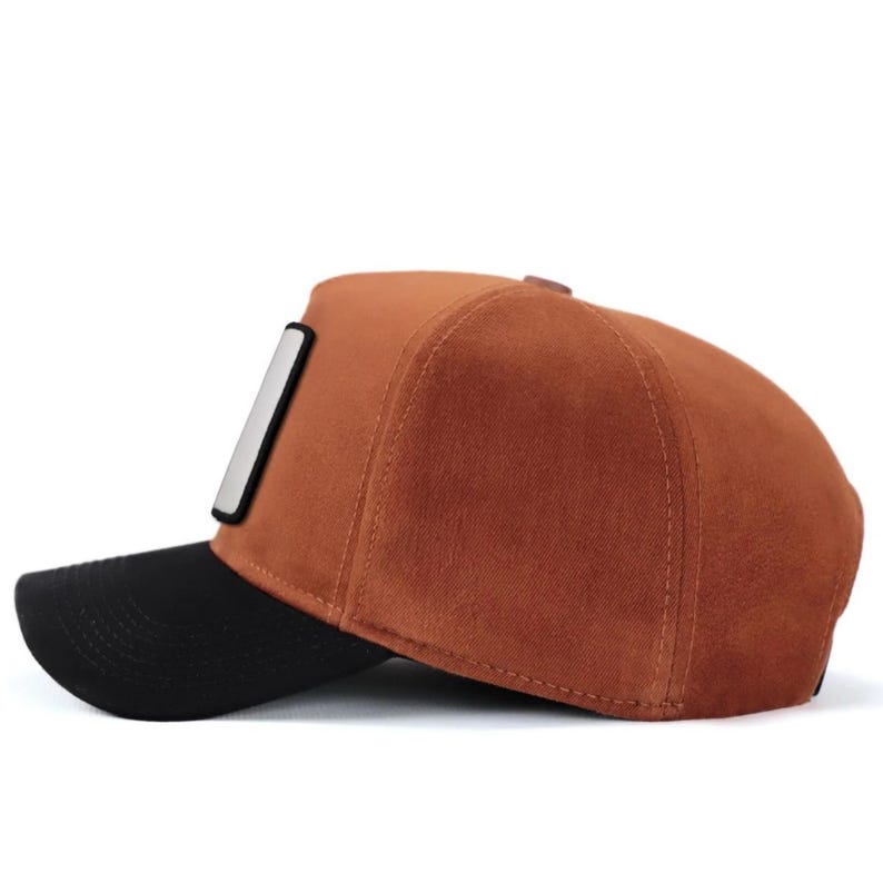 May include: A baseball cap featuring a brown suede-like crown and a solid black brim. The side of the cap displays a rectangular patch with a white center and a thick black border. The cap is constructed with visible stitching along the panels.