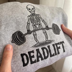 May include: A heather gray sweatshirt featuring black embroidery of a skeleton performing a deadlift with a barbell. The word DEADLIFT is embroidered in bold black capital letters below the skeleton graphic on the front of the garment.
