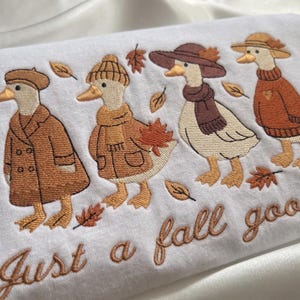 Pode incluir: Uma camisola branca com um design bordado de quatro gansos vestidos com roupas de outono, incluindo chapéus, cachecóis e casacos em tons de castanho e laranja, rodeados por folhas com o texto Just a fall goose bordado por baixo.