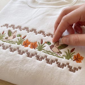 Könnte beinhalten: Ein weißes Sweatshirt mit Rundhalsausschnitt und einem horizontalen Stickmuster aus Stockenten, grünen Schilfhalmen, Rohrkolben und orangefarbenen Herbstblättern. Die Stickerei ist von zwei Reihen brauner geometrischer Stichmuster umrahmt.