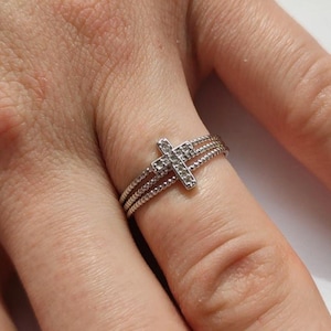 Könnte beinhalten: Silberfarbener Ring mit Kreuz-Design. Der Ring hat ein dreifaches Banddesign mit einem strukturierten, seilartigen Muster. Das Kreuz ist mit kleinen, funkelnden Steinen besetzt. Ein Schmuckstück für religiöse oder modische Zwecke.