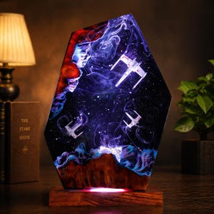 Peut inclure: Lampe géométrique unique représentant une scène spatiale avec des chasseurs stellaires blancs sur un fond de galaxie bleu foncé et violet. La lampe est en résine et en bois, avec une lueur rouge chaude émanant de la base.