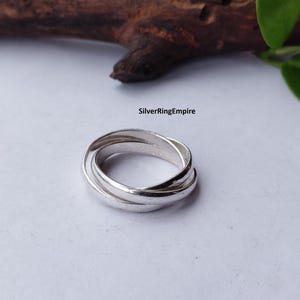 Può includere: Un anello color argento composto da tre fasce intrecciate che si sovrappongono per formare un'unità circolare. Il testo SilverRingEmpire è stampato in nero sopra l'anello. La superficie metallica è liscia, riflettente e lucida.