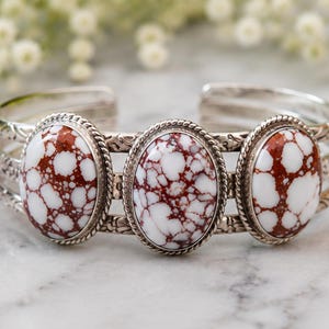 May include: A silver cuff bracelet featuring three oval-shaped stones with a white and reddish-brown webbed pattern. Each stone is set in a twisted rope border, and the silver band is decorated with an engraved floral pattern along the sides.