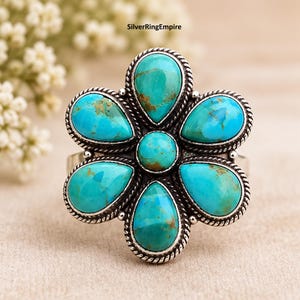 May include: A silver ring featuring a flower-shaped design with six teardrop-shaped turquoise stones surrounding a central round turquoise stone. Each stone is set in a twisted silver rope border. The text SilverRingEmpire is visible.