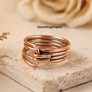 Puede incluir: Un anillo de color cobre con un diseño de múltiples bandas que presenta varios bucles interconectados que se superponen en la parte frontal. El metal tiene un acabado metálico cálido con sutiles variaciones de tono. El texto SilverRingEmpire es visible.