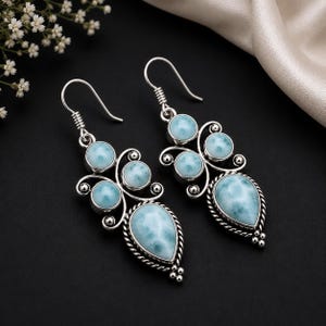 Puede incluir: Un par de pendientes colgantes de plata de ley con varias piedras preciosas Larimar de color azul claro en forma redonda y de pera. Los engastes de plata incluyen intrincados diseños de filigrana y detalles de cuerda trenzada.