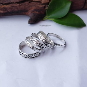 Puede incluir: Un conjunto de cuatro anillos de color plateado dispuestos en fila, cada uno con distintos patrones florales y geométricos en relieve. Los anillos tienen un acabado oxidado que resalta los detalles intrincados. El texto SilverRingEmpire es visible.