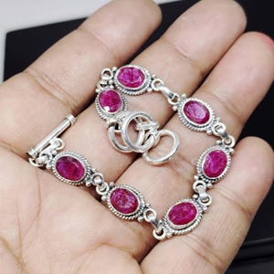Può includere: Un bracciale in argento con pietre preziose ovali rosso intenso. Le pietre sono incastonate in montature d'argento ornate e collegate da maglie d'argento. Il bracciale ha una chiusura a levetta. Il bracciale è esposto su una mano.