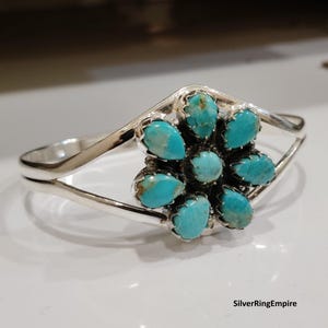 May include: A silver cuff bracelet featuring a central flower-shaped cluster of seven teardrop-shaped turquoise stones set in serrated bezels. The polished silver band splits into two arms that connect to the floral centrepiece. Text reads SilverRingEmpire.