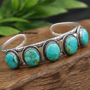 May include: A silver cuff bracelet featuring four oval turquoise stones with brown matrix patterns. The metal band has a textured, oxidised finish and twisted rope detailing around each stone setting. The text SilverRingEmpire is visible.