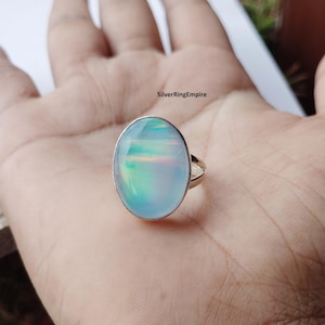May include: A silver ring featuring a large, oval-shaped, iridescent cabochon stone with a smooth, polished surface that displays soft blue and rainbow-like light reflections. The text SilverRingEmpire is visible above the ring.
