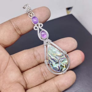 May include: A silver pendant featuring a teardrop-shaped abalone shell and two oval purple amethyst stones. The pendant is wire-wrapped and has a decorative bail. The abalone shell displays iridescent colours of green, blue, and gold.