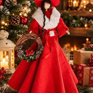 Puede incluir: Una muñeca decorativa con un vestido de fieltro rojo, cuello de piel sintética blanca y un sombrero puntiagudo rojo a juego. La muñeca tiene el pelo largo y oscuro y sostiene una pequeña corona circular de ramas, hojas y bayas rojas.