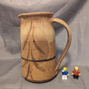 May include: A ceramic pitcher with a light tan upper section and a darker, speckled tan lower section. A dark horizontal line separates the two colors, and a stylized brown branch pattern is painted on the front. A curved handle is on the right side.