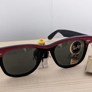 May include: Black sunglasses with a red patterned top frame and dark lenses. The sunglasses have a Ray-Ban logo sticker and a tag attached. The sunglasses are on a wooden surface.