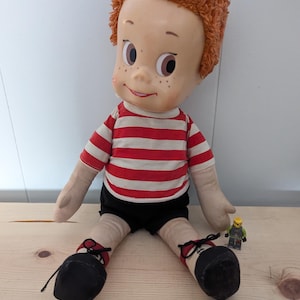 May include: A vintage doll with a painted face, freckles, and red hair. The doll wears a red and white striped shirt, black shorts, and black shoes with red accents. A small Lego figure sits nearby. The doll is approximately 20 inches tall.
