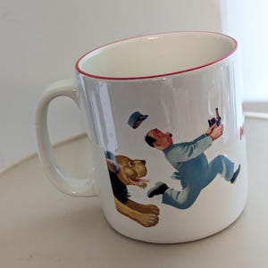 May include: White ceramic mug with a red rim and handle. The mug features a cartoon illustration of a man in blue overalls running from a dog. The man is holding a bottle. The mug has a white interior.