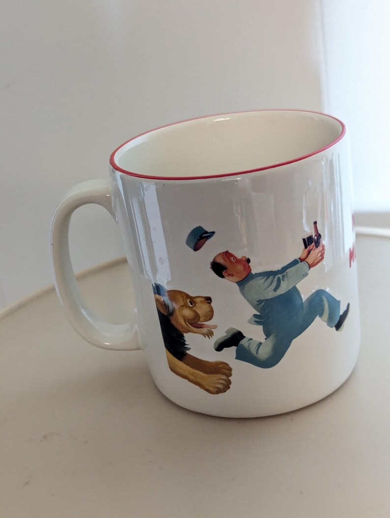 May include: White ceramic mug with a red rim and handle. The mug features a cartoon illustration of a man in blue overalls running from a dog. The man is holding a bottle. The mug has a white interior.