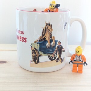 May include: White ceramic mug with a red rim, featuring a vintage illustration of a horse-drawn carriage and the text "Goodness Guinness". A Lego figure is inside the mug, and another is standing beside it.