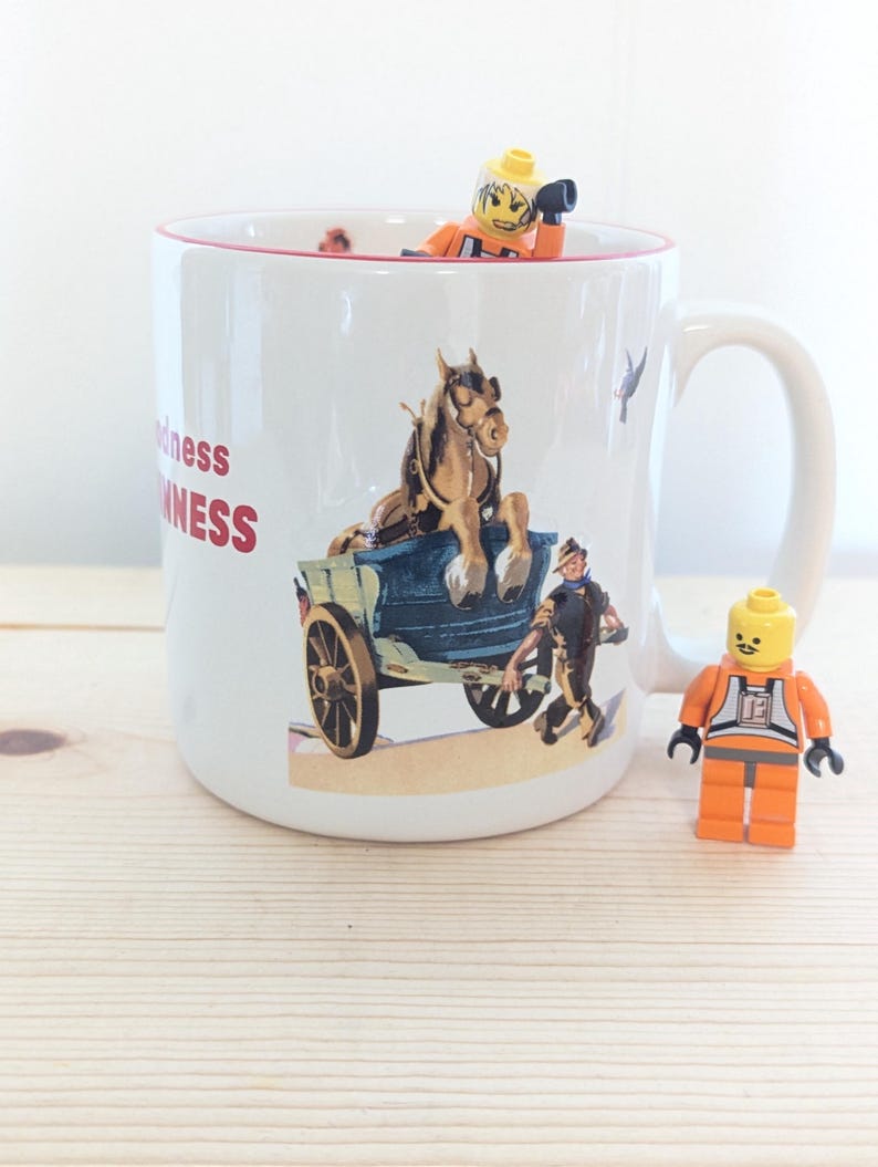 May include: White ceramic mug with a red rim, featuring a vintage illustration of a horse-drawn carriage and the text "Goodness Guinness". A Lego figure is inside the mug, and another is standing beside it.