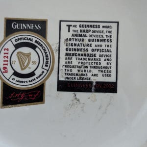 May include: Close-up of a white surface with two black and gold Guinness labels. One label is a circular official merchandise sticker with a harp logo and the text "St James's Gate Dublin". The other label contains text about Guinness trademarks.