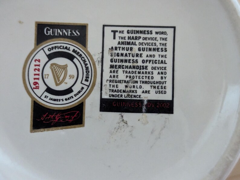 May include: Close-up of a white surface with two black and gold Guinness labels. One label is a circular official merchandise sticker with a harp logo and the text "St James's Gate Dublin". The other label contains text about Guinness trademarks.