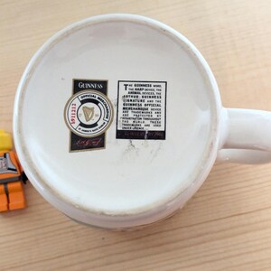 May include: White ceramic Guinness mug with a handle, viewed from the bottom. The mug has a black and gold Guinness Official Merchandise sticker. A small Lego figure in orange and yellow is next to the mug.
