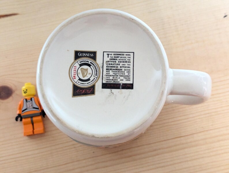 May include: White ceramic Guinness mug with a handle, viewed from the bottom. The mug has a black and gold Guinness Official Merchandise sticker. A small Lego figure in orange and yellow is next to the mug.
