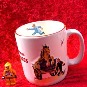 May include: White ceramic mug with a red rim and handle, featuring a vintage illustration of a horse-drawn carriage and a man running. The mug also has the word "mess" printed on it. A yellow Lego figure is in the image.