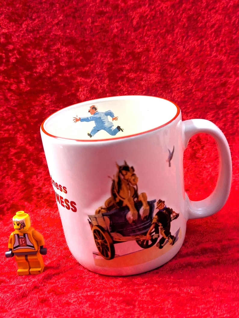 May include: White ceramic mug with a red rim and handle, featuring a vintage illustration of a horse-drawn carriage and a man running. The mug also has the word "mess" printed on it. A yellow Lego figure is in the image.