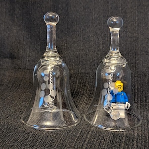 May include: Two clear glass bells with etched circular patterns on the exterior. Each bell features a silver-toned metal chain hanging from the interior top. One bell contains a small yellow-headed plastic toy figure wearing a blue shirt.