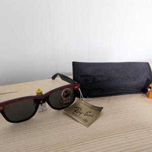 May include: Black Ray-Ban sunglasses with a red accent on the frame, a black case, and a tag. The sunglasses are displayed with two Lego figures on a light wooden surface.