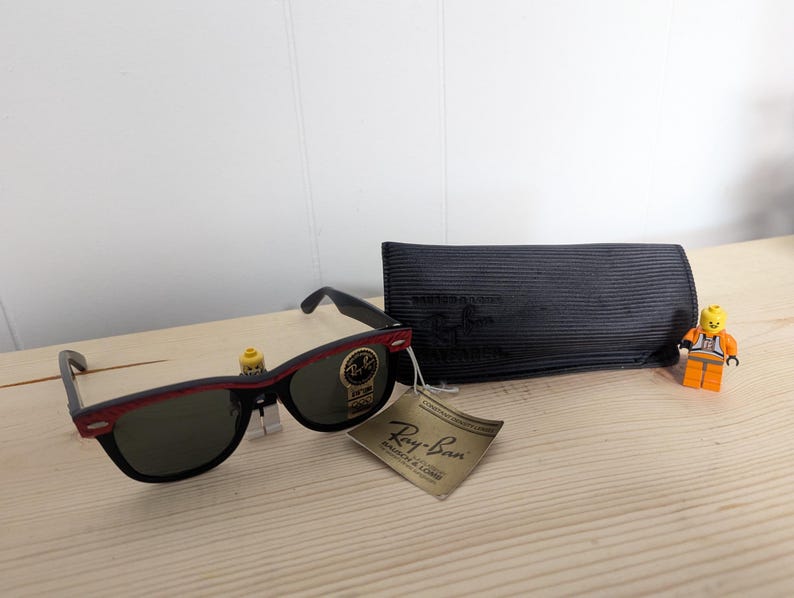 May include: Black Ray-Ban sunglasses with a red accent on the frame, a black case, and a tag. The sunglasses are displayed with two Lego figures on a light wooden surface.