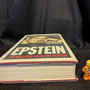 Puede incluir: Un libro titulado "Epstein: The Man Who Made The Beatles" de Ray Coleman, junto a una figura de Lego. El libro tiene una cubierta blanca con ribete rojo. La figura de Lego lleva un mono naranja.