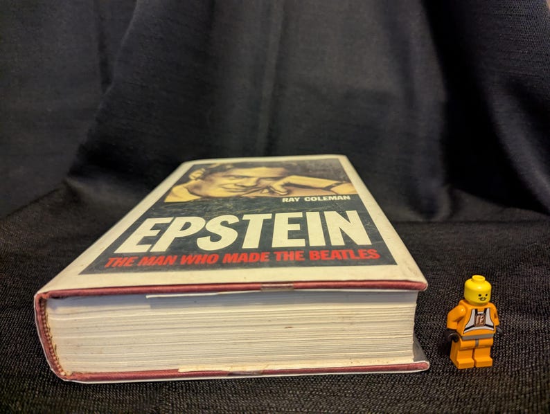 Puede incluir: Un libro titulado "Epstein: The Man Who Made The Beatles" de Ray Coleman, junto a una figura de Lego. El libro tiene una cubierta blanca con ribete rojo. La figura de Lego lleva un mono naranja.