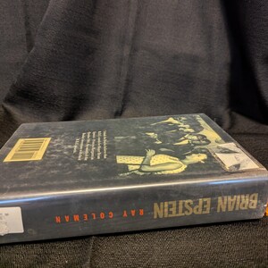 Puede incluir: Un libro de tapa dura titulado "Brian Epstein" de Ray Coleman. El libro tiene una cubierta oscura con una imagen en blanco y negro y texto en rojo y blanco. Una peque&ntilde;a figura de Lego naranja est&aacute; al lado del libro.