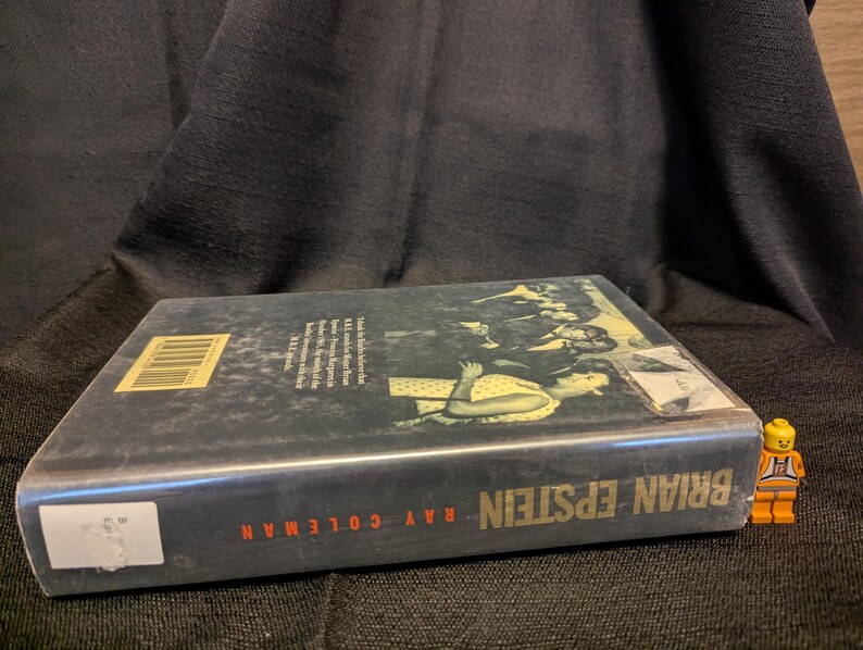 Puede incluir: Un libro de tapa dura titulado "Brian Epstein" de Ray Coleman. El libro tiene una cubierta oscura con una imagen en blanco y negro y texto en rojo y blanco. Una peque&ntilde;a figura de Lego naranja est&aacute; al lado del libro.